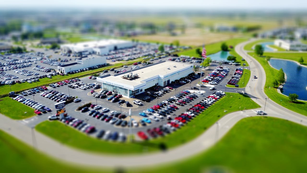 hero-img-01 Aerial high-angle view of a bustling car dealership surrounded by parked cars in a green landscape.